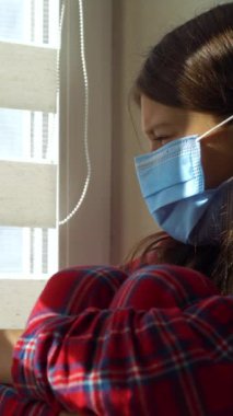 Dikey ekran: tıbbi maske ve pijama giyen bir kız pencerenin yanında oturuyor, dizlerine sarılıyor ve perdelerden bakıyor, üzerine güneş ışığı düşüyor. Çocuk salgın sırasında evde kalıyor. Sosyal uzaklık