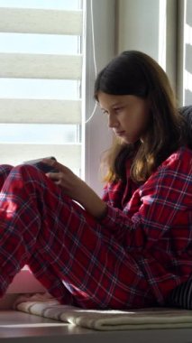 Pijamalı bir kız pencerenin yanındaki masada oturuyor ve akıllı telefondan online oyun oynuyor, güneş ışığı yüzüne vuruyor. Bağımlı çocuk evde kalıyor ve aletlerle vakit geçiriyor.