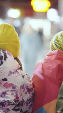 Kış giysileri içindeki çocuklar, şenlikli ışıklarla buz pateni pistinde durup bulanık insanların paten yapışını izliyorlar. Yılbaşı pazarında tanınmayan çocuklar. Tatil kavramı