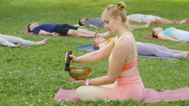 瞑想中にボウルを歌う若い女性は 実践者は死体のポーズに草の上に横たわっています シャヴァーナ ヨガクラスの深い修復 ボディー マインドリラクゼーションのコンセプトを実施する人々 — ストック動画
