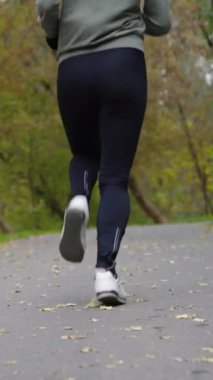 Spor kıyafetli formalı bir kadın yavaş çekimde yapraklarla yolda koşuyor. Sonbahar günü dışarıda aktif insan eğitimine kısmi bakış açısı. Spor anlayışı