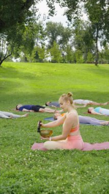 Dikey Ekran: kadın meditasyon sırasında müzik kaseleri çalarken pratisyenler çimlerin üzerinde ölü pozunda yatıyor. Shavasana 'da dinlenen insanlar, yoga sınıfı derin restorasyon, beden ve zihin rahatlığı.