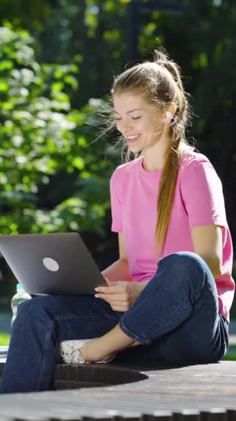 縦型スクリーン 晴れた日に公園のノートパソコンを介してビデオ通話を持つピンクのTシャツと青のジーンズを身に着けている若い女性 ジョギングや背景に赤ちゃんの馬車を持つ人々 オンラインコミュニケーションの概念 — ストック動画