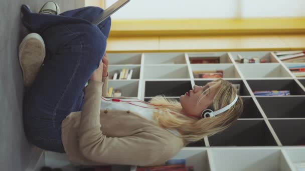 垂直スクリーン カジュアルな女性は 床に座ってノートパソコンで作業するヘッドフォンを着用し 本棚に傾いています リモートで働くフリーランサー インターネットを利用して図書館で学ぶ学生 — ストック動画