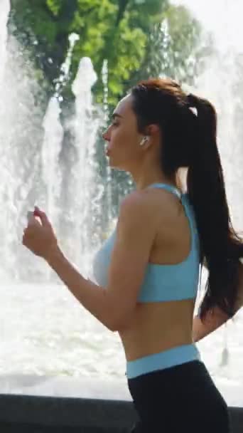 縦スクリーン 夏の噴水のまわりで走るスポーツ服およびイヤホンを身に着けているゆっくり動くフィットの女性 外で撮影された女性のジョギングトレーニング 健康的なライフスタイル フィットネスの概念 — ストック動画