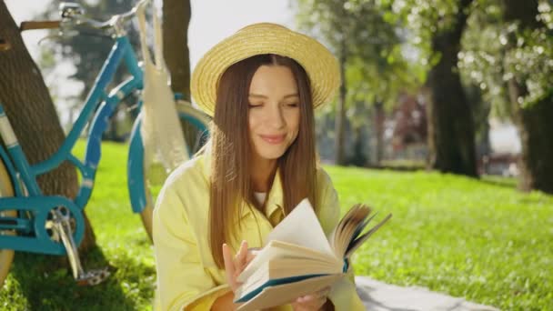 戴着草帽的年轻女子在绿色的草地上看书 蓝色的自行车在树旁 浪漫的女性眼睛紧闭 嘴角挂着微笑 梦幻般地向上凝视着 寻求孤独和灵感 — 图库视频影像