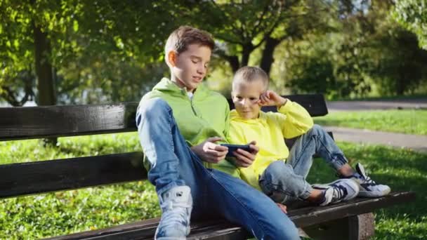 Старшие Младшие Братья Сидят Скамейке Солнечном Парке Смотрят Видео Смартфоне — стоковое видео