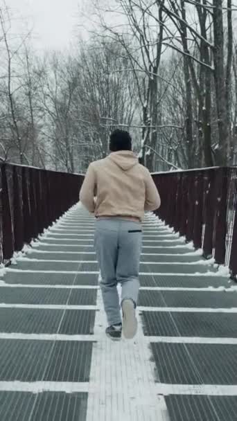 スローモーションで長い金属製の橋の上を走るスポーツウェアの男は 公園の下に横たわる雪で覆われている 冬にジョガーの訓練のショットの後 スポーツの概念 — ストック動画