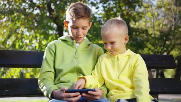 两个男孩坐在阳光灿烂的公园的长椅上 看着滑稽的视频 玩网络游戏 在夏日的室外用电话浏览互联网 和小玩艺儿在一起的闲暇时间吸毒成瘾的孩子技术概念 — 图库视频影像