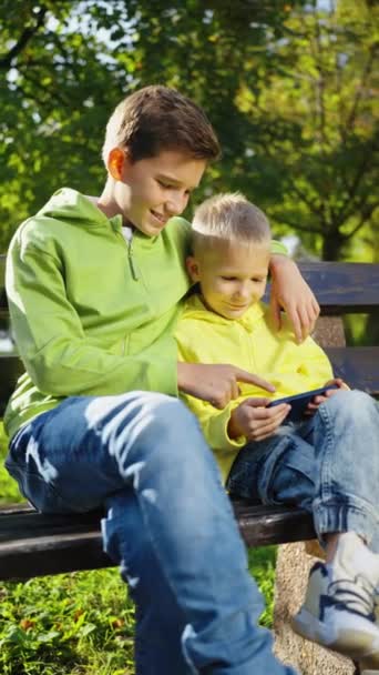 垂直屏风 两个男孩坐在公园的长椅上 观看滑稽视频 玩网络游戏 在室外浏览互联网 和小玩艺儿在一起的闲暇时间吸毒成瘾的孩子技术概念 — 图库视频影像