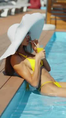 Havuzda meyve kokteyli içen şapkalı hoş bir kadın. Güneş gözlüklü, sarı bikinili, havuz kenarında içkili çekici bir kadın. Yaz tatili ya da tatil konsepti