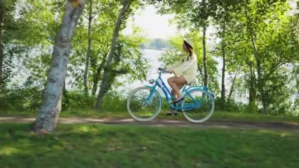 笑容可亲的女人戴着夏帽悠闲地骑着蓝色的自行车沿着公园的小径 背景模糊了河流 骑自行车的年轻女性感到快乐和放松 周末休闲 — 图库视频影像