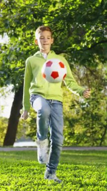 Dikey Ekran: Genç çocuk ayağıyla futbol topuyla hokkabazlık yapıyor. Güneşli parkta futbol antrenmanı yapan mutlu bir çocuk. Sağlıklı yaşam tarzı