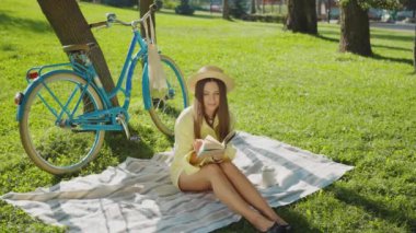 Hasır şapkalı romantik genç kadın yeşil çayır üzerine kitap okuyor, mavi bisiklet ağacın yanında. Neşeli bir kadın, kapalı gözlerle yukarı bakıyor ve dudaklarıyla gülümsüyor. Yalnızlık ve ilham arayışı