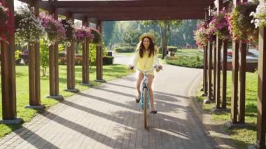 Yaz sabahı parkta mavi bisikletine binen sevimli genç bir kadın. Bisikletli güzel bir kadın çiçeklerin altından neşeyle geçiyor, ayaklarını pedallardan yana açıyor..