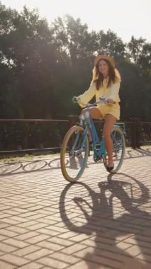 Yazlık şapkalı hoş bir kadın parkta mavi bisiklet sürüyor, uzun saçlarında güneş ışığı var. Bisikletli genç bir bayan mutlu ve rahat, hafta sonu tatili