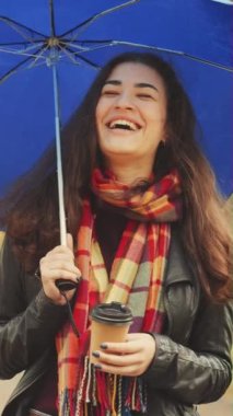 Dikey ekran: Sonbahar parkında kahve fincanı ve mavi şemsiyeyle gülen kareli atkı takan güzel mutlu genç bir kadın. Arka planda bulanık ağaçlar. Boş zaman kavramı.