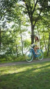 Dikey Ekran: Yaz şapkalı sevimli gülümseyen kadın parkta mavi bisiklet sürüyor, arka planda bulanık nehir. Bisikletli genç bir bayan mutlu ve rahat, hafta sonu tatili
