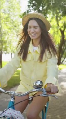 Dikey Ekran: Yazlık şapkalı sevimli gülümseyen kadın güneşli parkta mavi bisiklet sürüyor, saçlarında günbatımı ışınları var. Bisikletli genç bir bayan mutlu ve rahat, hafta sonu tatili