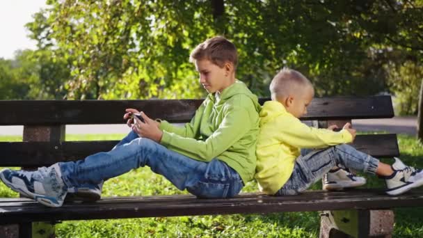 上瘾的电话用户 两兄弟背靠背坐在公园的长椅上 沉浸在手机里 而不是在玩积极的游戏 男孩们看有趣的视频 玩网络游戏 浏览互联网 — 图库视频影像