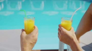 Oteldeki havuz başı şezlonglarında dinlenirken birbirine tokuşturan portakal suyu bardakları. Kadın ve erkek havuz kenarında içki içiyorlar. Tatil, rahatlama, dinlenme konsepti.