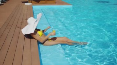 Şapkalı ve sarı bikinili tatlı bir kadın tatil köyünde meyve kokteyli içiyor. Güneş gözlüklü, havuz kenarında içecekle serinleyen çekici bir kadın. Yaz tatili ya da tatil konsepti