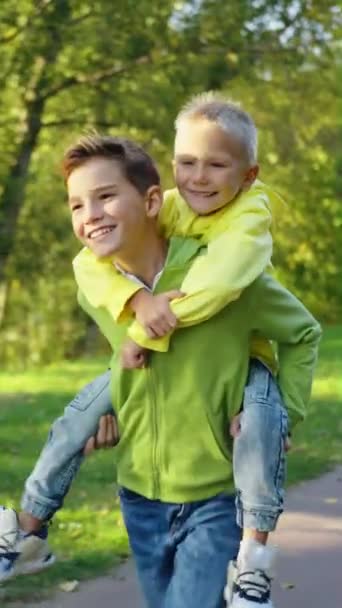 垂直屏风 快乐的孩子喜欢骑在哥哥的背上 笑着让小孩搭便车 男孩在阳光灿烂的公园里奔跑 快乐的孩子们玩得开心 — 图库视频影像