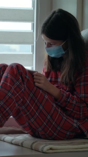 Vertikal Skärm Flicka Pyjamas Bär Medicinsk Mask Sitter Nära Fönstret — Stockvideo