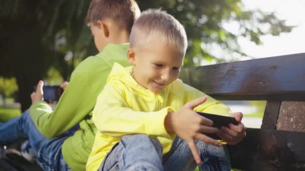 2人の幸せな笑顔の少年が ペアレンタルコントロールなしでオンラインで漫画を見ている携帯電話を閲覧します 公園のスマートフォンでゲームをする兄弟 現代のコミュニケーションとガジェット依存の概念 — ストック動画