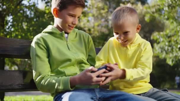 兄弟俩在公园的长椅上坐着玩电话时意见不一 年轻的男孩想从年长的男孩那里抢智能手机 行动成瘾概念 — 图库视频影像