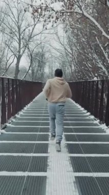 Spor giyimli ve şapkalı bir adam metal köprüde ağır çekimde koşuyor, yerde kar var. Kışın dışarıda atış koşucusu eğitimini takip ediyorum. Spor anlayışı