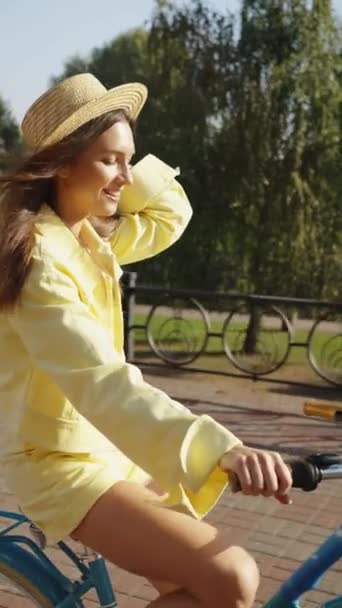 垂直屏风 快乐的年轻女子在早上的城市公园骑自行车 女人们紧紧抓住她的夏帽以防止风吹走它 感到快乐和放松 周末休闲 — 图库视频影像