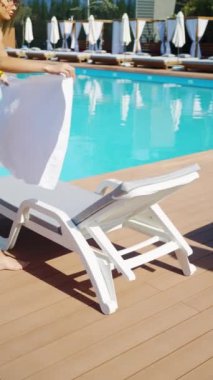 Sarı bikinili genç bir kadın havuz kenarındaki plaj salonuna beyaz havlu seriyor. Büyüleyici bayan güneşleniyor ve otelde dinleniyor.