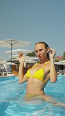 Güneş gözlüklü ve yüzme havuzunda poz veren sarı bikinili genç bir kadın. Havuzda kristal suyla kameraya flört eden güzel bir kadın portresi. Yaz ve tatil, güneş banyosu, yaşam tarzı konsepti