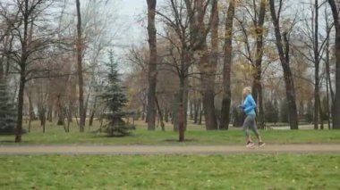 Yetişkin bir kadın sonbahar parkında koşuyor, mavi ceket ve gri taytlar giyiyor çıplak ağaçların arasında. Tuğla yolda koşan, yeşil çimenlerin arasından dökülen yapraklarla örülmüş sportif bir dişi..