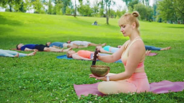 女性は 静かな屋外瞑想でサウンドセラピーを結合し 街の公園でマットに休むようにリラクゼーションでリラックスして歌うボウルを果たしています — ストック動画