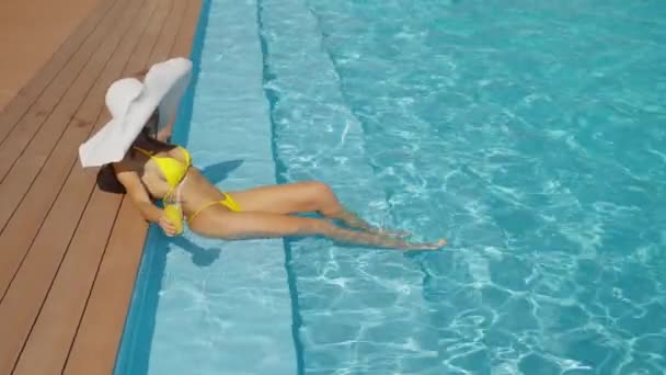身穿黄色比基尼和白色大太阳帽的优雅女人躺在游泳池边 双腿浸在水里 手里拿着柑橘饮料 快乐的女性在豪华的游泳池边放松 假期的概念 — 图库视频影像