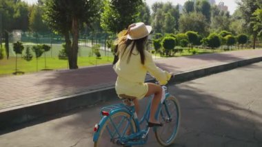 Sarı elbiseli kadın, güneşli bir günde hasır şapkasını tutarak park yolu boyunca bisiklet sürmekten zevk alıyor. Mutlu ve rahatlamış hissediyorum, hafta sonu tatili.