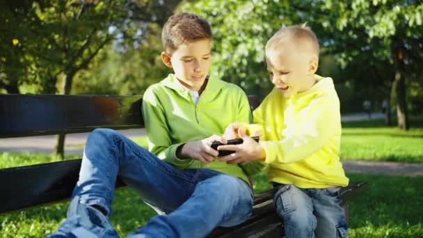 两个穿着鲜艳羊毛衫的兄弟一边坐在公园的长椅上 一边开玩笑地争夺着智能手机的所有权 一边捕捉着兄弟间的竞争和乐趣 — 图库视频影像