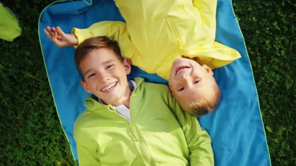 两个快乐的兄弟一边笑着一边在茂盛的野餐毛毯上放松着 四周是茂密的绿叶 笑着的兄弟姐妹们捕捉着纯粹的快乐和联系的时刻 — 图库视频影像