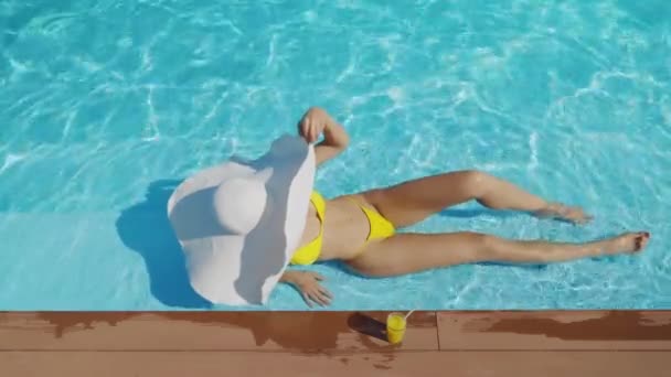 身穿黄色比基尼的女人躺在游泳池边 手里拿着一顶宽边帽 旁边还有一杯清爽的饮料 迷人的女性日光浴和休闲在度假酒店 — 图库视频影像