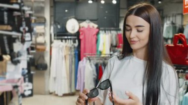 Moda mağazasında şık güneş gözlüğü deneyen neşeli bir kadın. Moda kıyafetleriyle çevrili. Bayan müşteri tatil için aksesuar alıyor, kameraya bakıyor ve gülümsüyor. Alışveriş kavramı