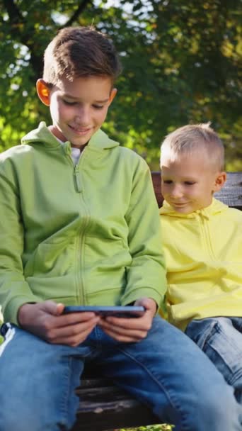 哥哥和弟弟坐在阳光充足的公园的长椅上 在智能手机上观看视频 两个男孩在电话上花时间 而不是在玩积极的游戏 行动成瘾概念 — 图库视频影像