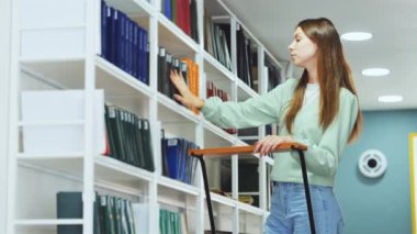 Genç kadın kitaplıkları karıştırıyor, kütüphanede çalışma materyali seçiyor, sahne bilgi arayışını resmediyor. Akademik araştırma ve bilgi arayışı