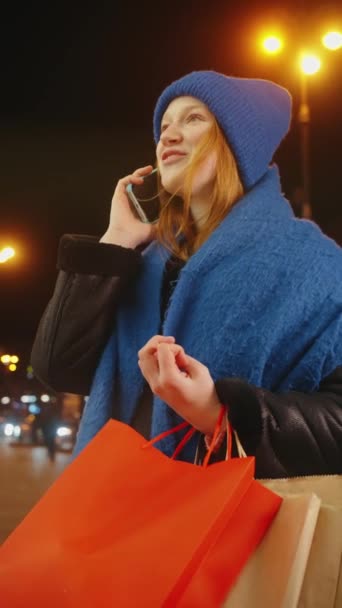 楽しい女性は彼女の携帯電話でチャット 夜に活気のある通りでショッピングバッグを運ぶ 彼女の冬の服を照らす街灯の暖かい光 — ストック動画