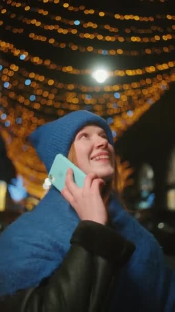 ツインリングライトの下で電話中に笑顔の女性 青い帽子の若い女性は 夜の背景でボケ効果を作成するお祝いのライトで電話中に喜んで笑います — ストック動画