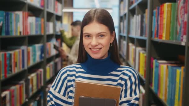 Joyful Young Woman Striped Sweater Smiling Camera Holding Notebooks Bookshelves — Stock Video