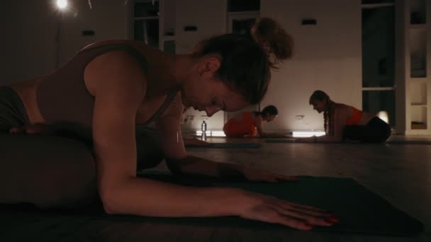 年轻女子练习静坐瑜伽时 在灯光昏暗 气氛轻松的房间里摆姿势 其他人背景在垫子上 流露出宁静和专注 健康与团体健身概念 — 图库视频影像