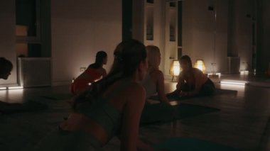 Kadın pratisyenler sıcak ışıklandırılmış stüdyoda esneme hareketleri yaparak sakin bir grup ortamı yaratırlar. Yoga dersine katılan kadınlar loş ışıklı bir odada, duvarda hafif ışıklandırma ve aynalar var.