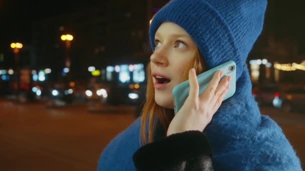身着蓝色针织帽子的年轻女子 深夜在繁忙的城市街道上用智能手机交谈 背后的红绿灯模糊不清 — 图库视频影像